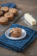 Homemade Blueberry Muffins on Wooden Table Top; One Isolated on White Plate on Blue Kitchen Towel; More Muffins on Cooling Rack in Background; Butter Dish and Knife beside the Muffin 