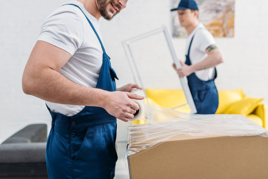 cropped view of mover in uniform wrapping cardboard box with stretch film in apartment