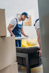 selective focus of movers wrapping couch with roll of stretch film in apartment