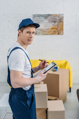 handsome mover holding clipboard and looking at camera near cardboard boxes in apartment