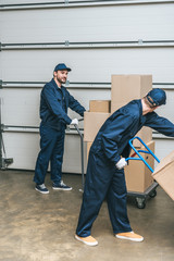 Fototapeta premium two movers in uniform using hand trucks while transporting cardboard boxes in warehouse