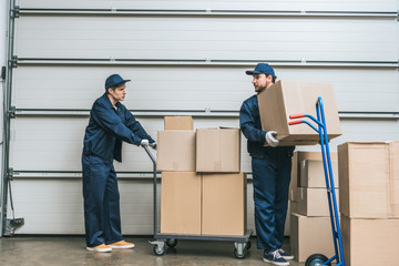 two movers in uniform looking at each other while transporting cardboard boxes with hand trucks in warehouse