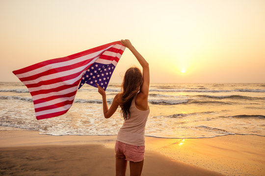 view from the back young brunette female person holding national American flag cover ocean beach holiday travel at summer romantic sunset .America independence day concept