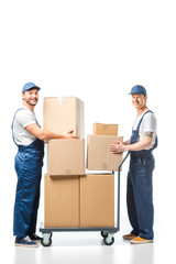 two handsome movers looking at camera while transporting cardboard boxes with hand truck on white