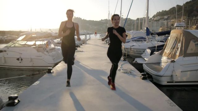 Young women doing exercises at sea port. Fit attractive caucasian girls run along the pier between the yachts