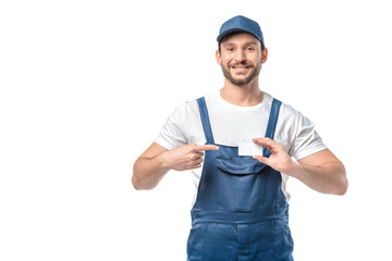 smiling mover in uniform looking at camera and pointing with finger at blank business card isolated on white