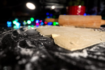 Uncooked rolled dough on table in kitchen. Christmas or fairy lights and decorations in background. Flour all around. Baking gingerbread, cookies, bread or pastry