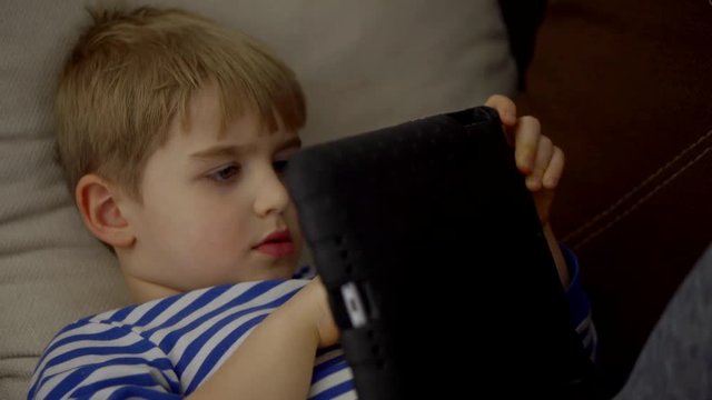 European baby boy sitting in bed, playing with tablet PC. Children playing with tablet computer, gadget-dependent children, the concept of pseudo-autism