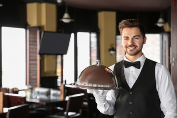 Young male waiter with tray and cloche in restaurant