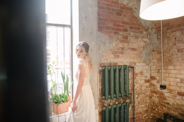Model posing in a white long wedding dress indoors by the window. Advertising of wedding dresses