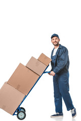 smiling handsome mover in uniform transporting cardboard boxes on hand truck isolated on white