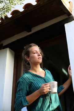 Young woman with cup of coffee standing at house entrance looking at distance