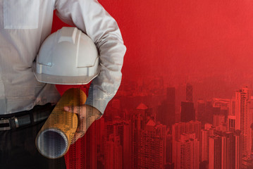 Double exposure of male civil engineer holding protective safety helmet and architectural drawing near red concrete wall with Hong Kong city background. Architecture design and construction business