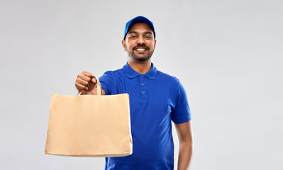 service and people concept - happy indian delivery man food in paper bag in blue uniform over grey...