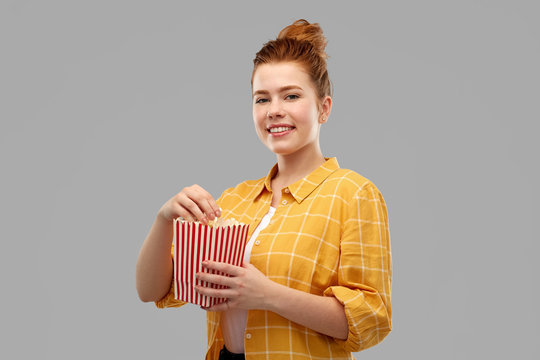 fast food and people concept - smiling red haired teenage girl in checkered shirt eating popcorn from striped bucket over grey background