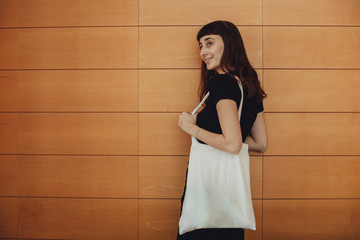 Young hipster smiling girl with white blank cotton shopper tote bag wearing black t-shirt and black jeans, mock-up of blank bag, wooden wall on the background, concept of shopping