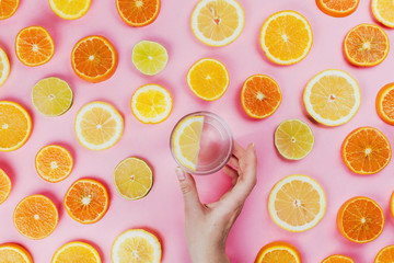 Flatlay of woman's hand holding lemon water on pink background with various sliced citrus fruits