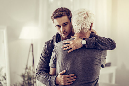 Twenty-years-old dark-haired kind male calmingly cuddling sick sorrowful aging granddad