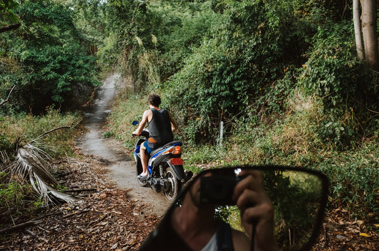 Young man on a scooter driving on a jungle path while another man can be seen through the rear mirror taking a picture of him