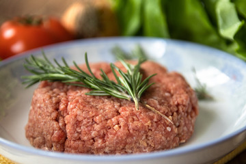 Fresh raw minced meat with rosemary on a plate and tomato, garlic, lettuce and onion on wooden table. Composition with meat and fresh organic vegetable.