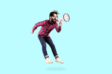 Bearded hipster man in glasses, jumping and shouting in megaphone, photo on blue background