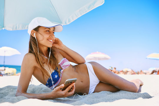 beautiful tanned happy laughing woman is lying on the beach on a white sand, relaxing and listen music from the phone with small headphones. slim sexy woman in bikini and white cap enjoing and lying