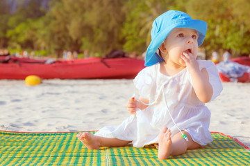 Little baby girl 8 month is sitting on the sandy beach in blue hat and white dress. She is impressed of the sea.