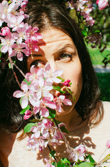 Fototapeta premium brunette girl stands near a blossoming branch of a Apple tree. pink flowers on the face. it's hard to look at the sun, hurts my eyes
