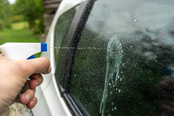 A hand holding a plastic spray bottle splashes onto a car's glass.