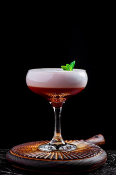 Elegant red ladies' cocktail with pink foam in a beautiful glass on a wooden carved round board. Cocktail isolated on black background. Close-up. Space
