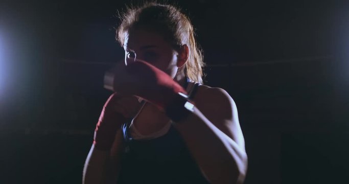 A beautiful female boxer strikes directly into the camera looking into the camera and moving forward on a dark background with a backlight. Steadicam shot