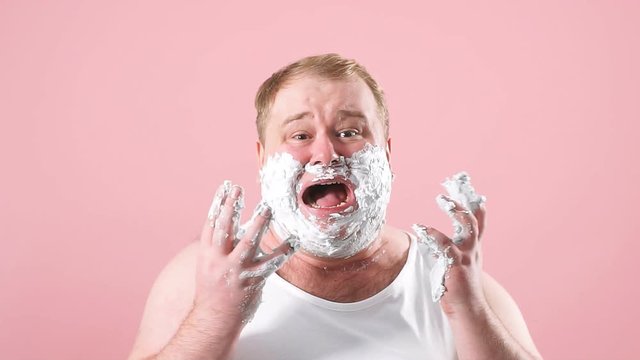 Upset man with gel on cheeks, has sad expression, sensitive skin, man going to shave his chin despite ofskin irritation, isolated over pink background.
