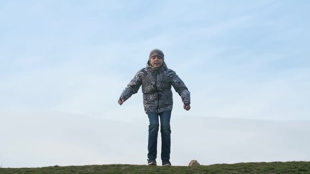 Happy boy in a jacket sitting and jumping up in a lawn on a sunny day in slo-mo 