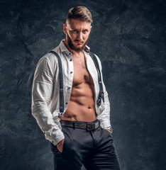 A stylish young man with a muscular body in the unbuttoned white shirt with suspenders. Studio...