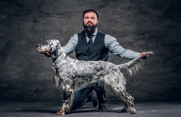 Middle-aged hunter dressed in elegant clothes sits on his knee and shows the full length of his purebred English Setter. Studio photo against a dark textured wall - obrazy, fototapety, plakaty