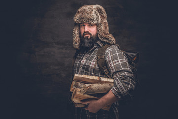 Portrait of a bearded woodcutter with a backpack dressed in a plaid shirt and trapper hat holding...