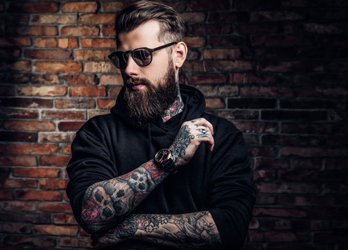 A stylish tattooed guy in a black hoodie and sunglasses. Studio photo against a brick wall