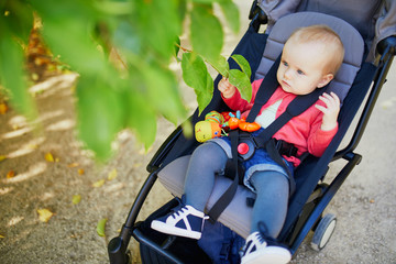 Adorable baby girl sitting in stroller and looking at leaves on a fall or spring day in park