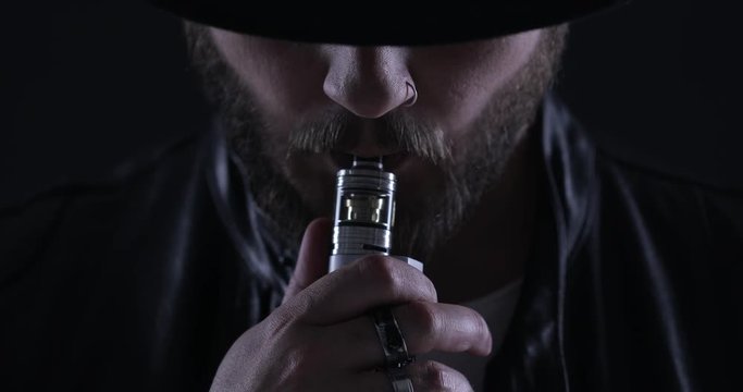 Man with vaping mod exhaling steam at black studio background. Bearded guy smoking e-cigarette to quit tobacco. Vapor and alternative nicotine free smoking concept