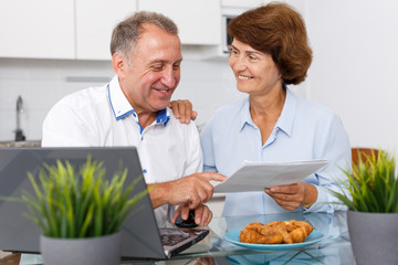 Fototapeta na wymiar Smiling mature family couple with documents using laptop at kitchen table