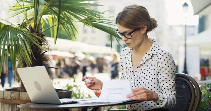 Pretty woman in the glasses having a lunch in the restaurant terrace and working at the laptop and documents. Outdoor.