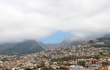 Fototapeta na wymiar a scenic panoramic aerial cityscape of the city of funchal in Madeira with buildings of the city in front of mountains with the tops covered by white clouds