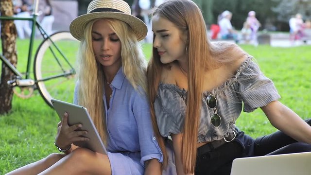 Two sociable girlfriends surfing the web laughing at fun pictures on the tablet sitting outdoors in the park in sunny weather.