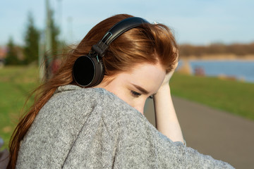 Young, sad cry redhead girl in the spring in the park near the river listens to music through wireless bluetooth headphones