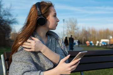 Young, happy redhead girl in the spring in the park near the river listens to music through wireless bluetooth headphones