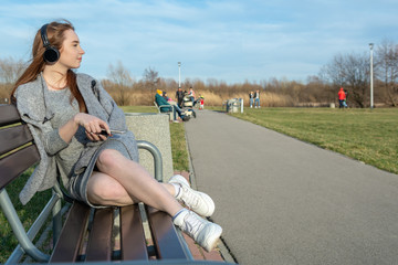 Young, happy redhead girl in the spring in the park near the river listens to music through wireless bluetooth headphones
