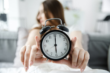 Tired young woman is holding an alarm clock in outstretched hands. Summer time concept.