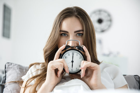 A young woman is holding an alarm clock with her hands, she is mysteriously looking at the camera. Daylight saving time concept.