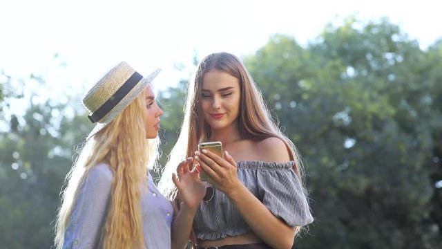 Cheerful girlfriends talking and browsing on smartphone on wonderful sunny summer day in the park.