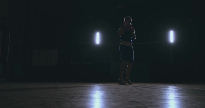 A beautiful woman boxer trains in a dark gym and works out punches in slow motion. Camera movement side View. Steadicam shot